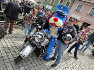 Zdjęcie kolorowe: policyjna maskotka na motocyklu, obok stoi kobieta i mężczyzna