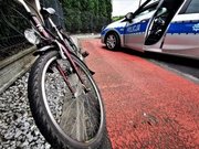 Zdjęcie kolorowe: z lewej strony widoczne koło przednie z roweru, w tle oznakowany policyjny radiowóz.