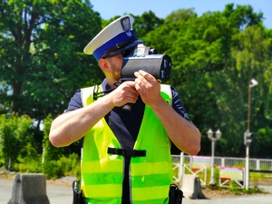 Zdjęcie kolorowe: umundurowany policjant ruchu drogowego podczas pomiaru prędkości.