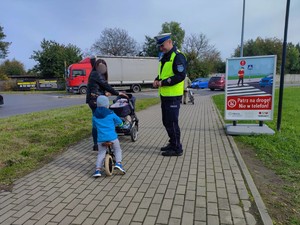 Zdjęcie kolorowe przedstawia umundurowanego policjanta, która uśmiecha się do chłopca na rowerku. Obok dziecka stoi kobieta z wózkiem.