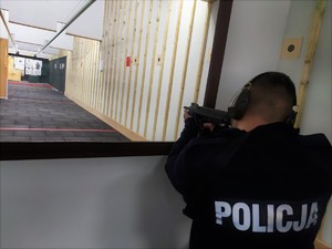 Zdjęcie przedstawia policjanta, który strzela kucając z broni maszynowej.