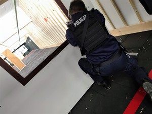 Zdjęcie przedstawia policjanta, który strzela z broni maszynowej zza przesłony, klęcząc na jednym kolanie.
