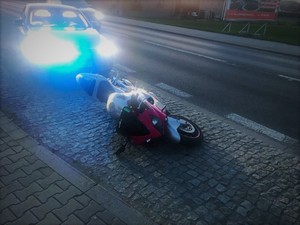 Zdjęcie przedstawia motocykl, który leży na ziemi, za nim zaparkowany samochód osobowy z włączonymi sygnałami świetlnymi.
