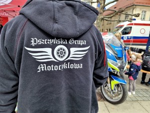 Zdjęcie przedstawia: tył czarnej bluzy, na której widnieje napis &quot;Pszczyńska grupa motocyklowa&quot;. Z tyłu widoczny policyjny motocykl.