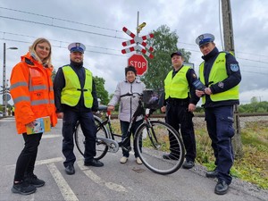 Zdjęcie przedstawia: kobietę w odblaskowej kurtce, policjanta, starszą kobietę z rowerem, funkcjonariusza Straży Ochrony Kolei i policjanta. Z tyłu przejazd kolejowy.