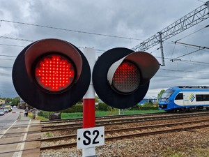 Zdjęcie przedstawia: sygnalizator świetlny w cyklu nadania światła czerwonego, z tyłu widoczny nadjeżdżający pociąg.
