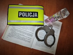 Zdjęcie przedstawia otwartą książkę, a na niej policyjne kajdanki i opaska z napisem Policja, obok stoi perfum.