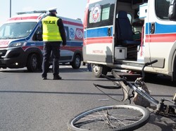Zdjęcie przedstawia uszkodzony rower, z tyłu karetka, umundurowany policjant.