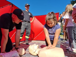 Zdjęcie przedstawia: dziewczynkę, która ćwiczy masaż serca, obok strażacy.