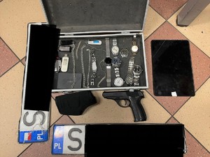 Zdjęcie przedstawia: walizkę w której są m.in. zegarki. Obok leży przedmiot przypominający broń i kabura oraz dwie tablice rejestracyjne.