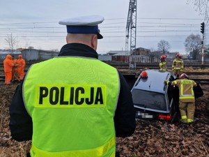 Zdjęcie przedstawia: umundurowany policjant, z tyłu widoczny samochód po zdarzeniu drogowym.