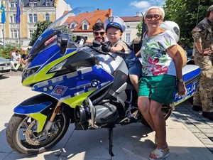 Zdjęcie przedstawia dwie osoby dorosłe i dziecko, które pozują do zdjęcia przy policyjnym motocyklu.