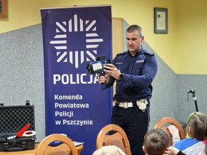 Zdjęcie przedstawia: 
Funkcjonariusza policji tłumaczącego przedszkolakom jak działa przyrząd do pomiaru prędkości.