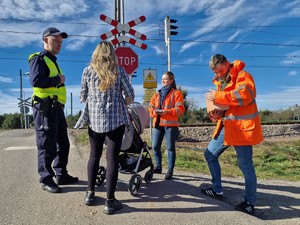 Zdjęcie przedstawia: 
Kobietę z wózkiem dziecięcym rozmawiającą z przedstawicielami Kolei Śląskiej i funkcjonariuszem policji, stojących przed przejazdem kolejowym.