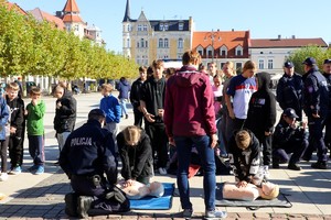 Na zdjęciu umundurowany policjant wraz z dziećmi ćwiczącymi prowadzenie resuscytację na fantomie