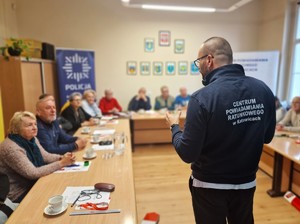 Zdjęcie przedstawia: mężczyzna w bluzie z napisem &quot;Centrum Powiadamiania Ratunkowego w Katowicach&quot; przed grupą seniorów.