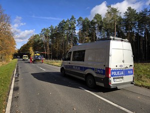Zdjęcie przedstawia oznakowany radiowóz na miejscu zdarzenia, w tle karetka oraz helikopter Lotniczego Pogotowia Ratunkowego.