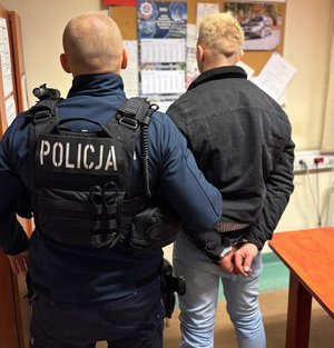 na zdjęciu zatrzymany w kajdankach stoi tyłem z policjantem