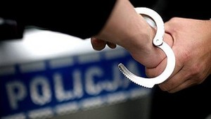 na zdjęciu w tle widoczny radiowóz oraz kajdanki zakładane na dłonie