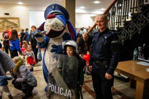 zdjęcie przedstawia policjanta, dziecko i maskotkę pasa Sznupka
