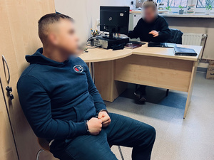 zdjęcie w pomieszczeniu biurowym, zatrzymany sprawca siedzi na krześle z kajdankami na rękach, z tyłu widać biurko przy którym siedzi przesłuchujący policjant