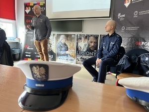 zdjęcie w klasie lekcyjnej przedstawiające czapki policjantów z drogówki leżące na stoliku, w tle policjanci i kierowca rajdowy