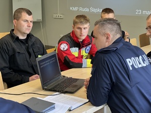 zdjęcie - policjant przy laptopie z tyłu trzej strażacy na sali odpraw