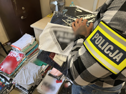 zdjęcie - w pomieszczeniu biurowym rozłożone zarekwirowane ubrania, policjant trzyma kilka bluzek w rękach
