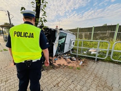 zdjęcie z miejsca zdarzenia - leżący na boku samochód dostawczy, obok drzewo w które uderzył, stojący przy nim policjant