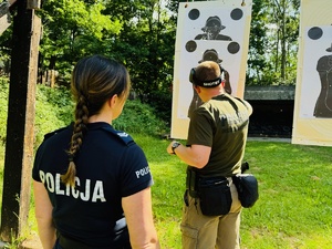 zdjęcie przedstawia zajęcia na strzelnicy otwartej podczas których instruktor ocenia wyniki strzelania na tarczy, przed stoi przyglądająca się policjantka