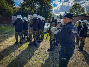 zdjęcie przedstawia szkolenie strażników granicznych - policjant wydaje komendy dla stojących czwórkami strażników wyposażonych w tarcze, hełmy i kamizelki