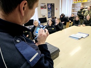 zdjęcie przedstawia uczniów klasy mundurowej przyglądającym się pokazywanemu przez policjanta miernikowi prędkości z funkcją nagrywania wykroczeń