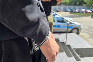 zdjęcie przedstawia ręce zatrzymanego z kajdankami na tle policyjnego radiowozu