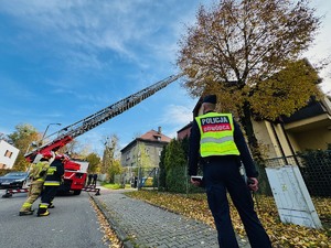 zdjęcie przedstawia policjanta, strażaków i wóz drabiniasty przy komisariacie