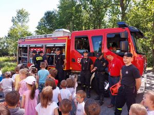 dzieci stoją przy wozie strażackim, przy którym stoją strażacy