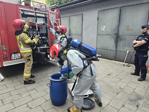 na zdjeciu strażacy wkładają do beczki pojemnik chemiczny