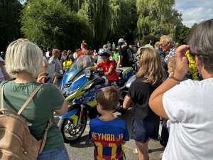 Zdjęcie przedstawia grupę ludzi. W tle dziecko na motocyklu służbowym.