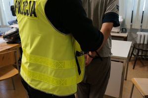 Zdjęcie przedstawia mężczyznę w kajdankach założonych na ręce trzymane z tyłu i policjanta w kamizelce z napisem &quot;Policja&quot;.