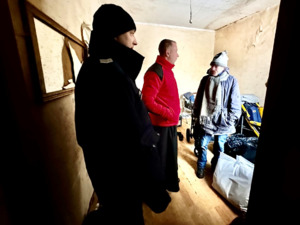 Zdjęcie przedstawia umundurowanych policjantów oraz księdza odwiedzających osobę potrzebującą w pomieszczeniu.
