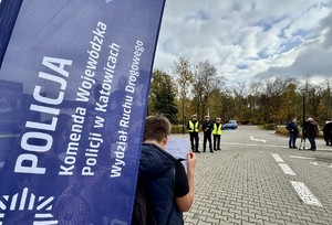 Zdjęcie przedstawia napis &quot;Policja Komenda Wojewódzka Policji w Katowicach Wydział Ruchu Drogowego&quot;. W tle trzech, umundurowanych policjantów.