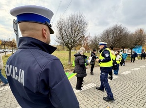 Zdjęcie przedstawia dwóch umundurowanych policjantów. Jeden z nich wręcza ludziom elementy odblaskowe.