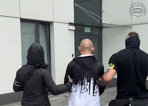 Zdjęcie przedstawia nieumundurowanych policjantów prowadzących osobę zatrzymaną do jednostki policji.