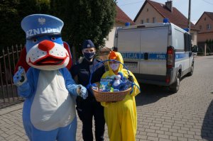 Na zdjęciu mundurowa wraz z maskotkami - Sznupkiem i Wielkanocnym Kurczakiem który trzyma koszyk z upominkami. W tle policyjny radiowóz.