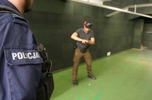 Na zdjęciu instruktor wyszkolenia strzeleckiego ze świętochłowickiej komendy, który trzyma w dłoniach broń i pokazuje policjantowi sposób sprawdzania broni po oddaniu strzału do celu, po lewej stronie widać napis policja na mundurze policjanta