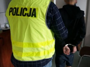 Na zdjęciu nieumundurowany policjant w żółtej kamizelce z napisem Policja, prowadzi zatrzymanego mężczyznę zakutego w kajdanki