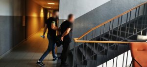 Na zdjęciu neumundurowany policjant z opaska odblaskową z napisem policja na prawym ramieniu prowadzi zatrzymanego mężczyznę zakutego w kajdanki na ręce trzemane z tyłu. Mundurowi prowadzi mężczyznę po schodach na wyższa kondygnację budynku
