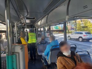 Na zdjęciu widać policjanta z drogówki wewnątrz autobusu w trakcie przeprowadzenia kontroli w związku z obowiązkiem zakrywania ust i nosa. Na zdjęciu widać siedzących paseżarów w maseczekach na twarzy