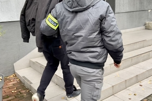nieumundurowany policjant trzymający mężczyznę z kajdankami założonymi za plecami wchodzący do budynku komendy