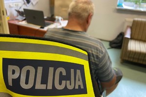 zdjęcie przedstawia opaskę z napisem policja a w tle siedzący mężczyzna