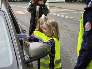 zdjęcie przedstawia policjantów z dziećmi ubranymi w kamizelki odblaskowe podczas akcji profilaktycznej wręczające laurki kierowcom